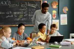 A teacher standing behind children sitting at a table looking at laptops.